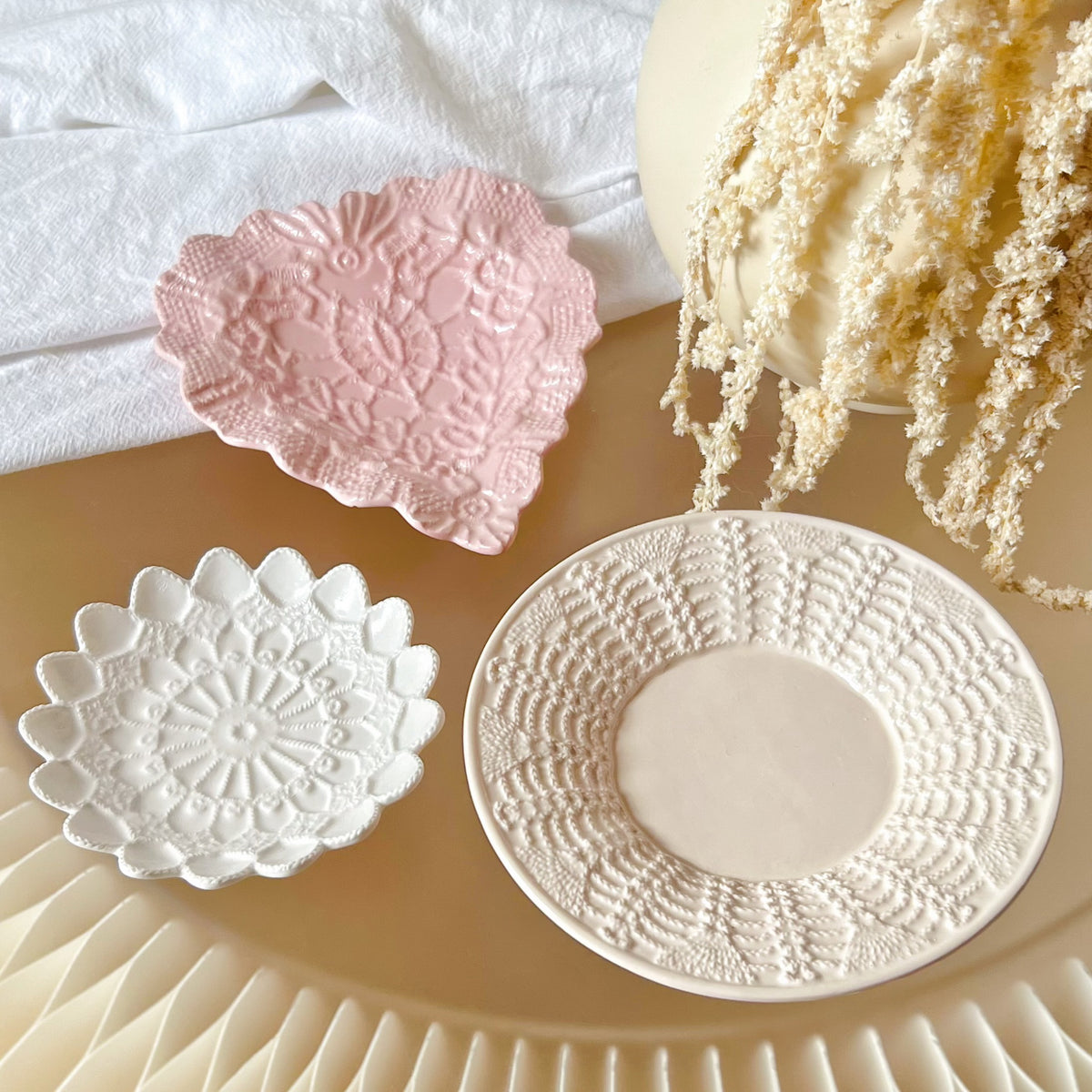 Three handmade decorative plates from LMJ Candles with embossed floral and geometric lace-like patterns, shown in white and soft pink on a warm beige surface with styled props including a white cloth and dried cream flowers.