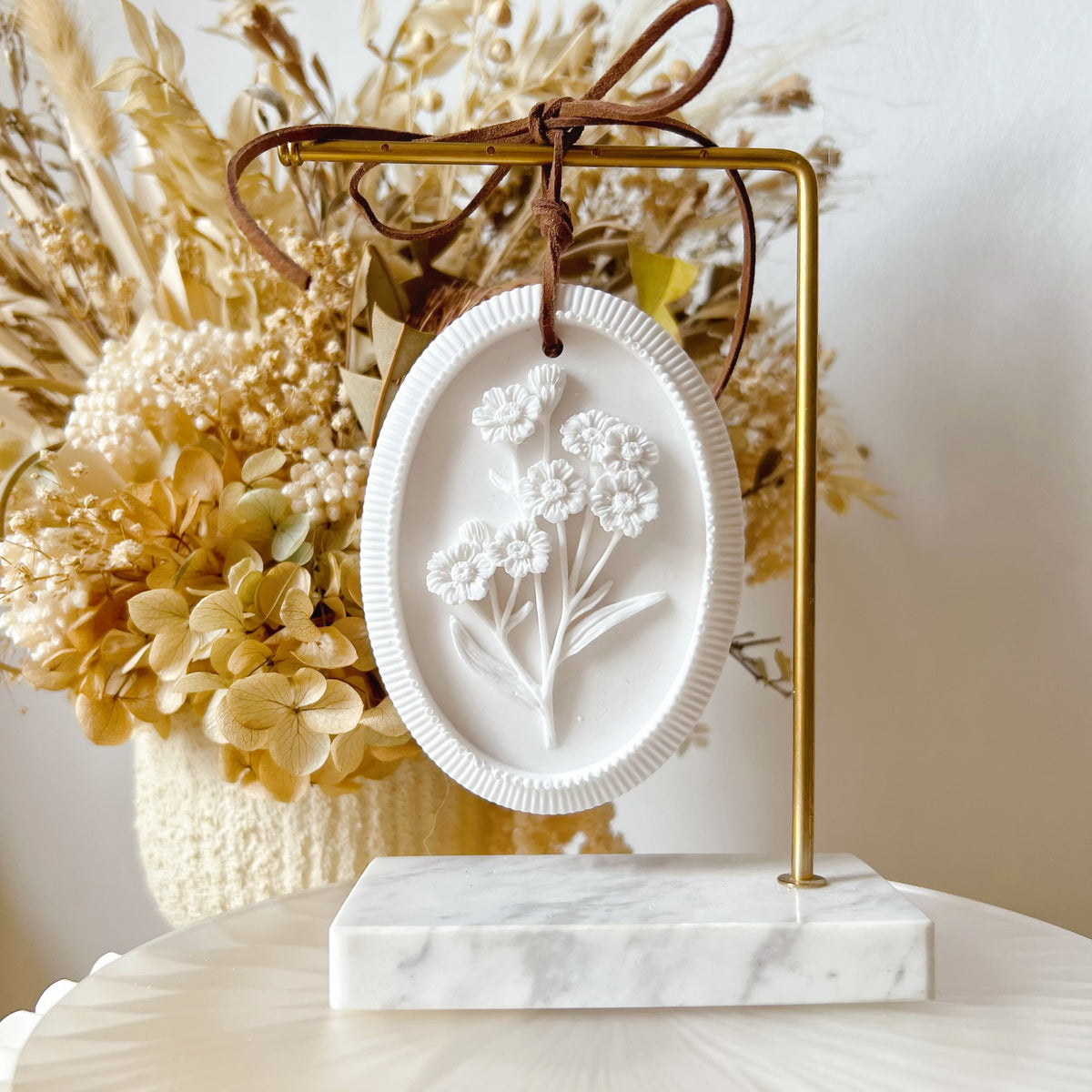 A white oval wall plaque car air freshener from LMJ Candles with a raised floral design, hanging from a gold metal stand with a brown ribbon, set on a marble-topped surface with dried flowers in the background.