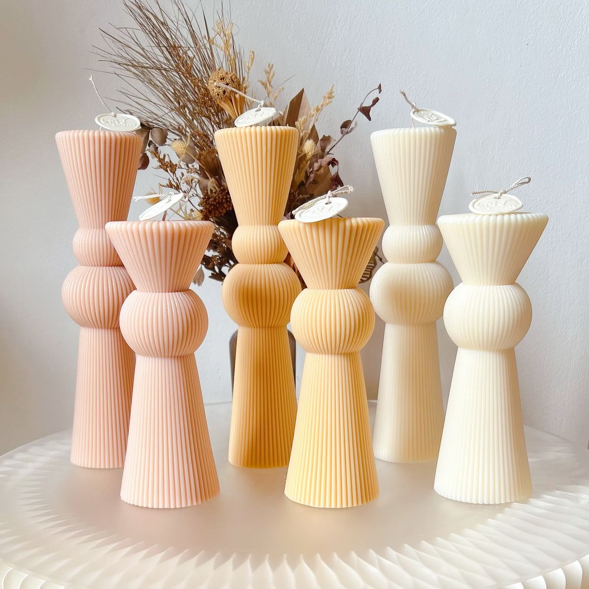 Six ribbed pillar candles from LMJ Candles in soft pastel shades of peach, orange, and cream, arranged on a white textured surface with a dried floral arrangement in the background.