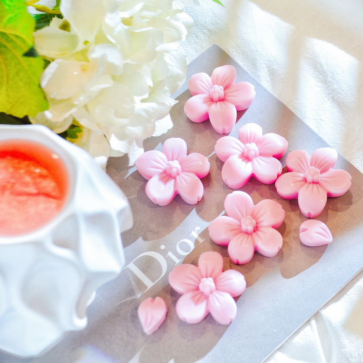 Pink cherry blossom flower soy wax melts from LMJ Candles arranged on a gray book next to a white ceramic cup with pink liquid and white flowers.